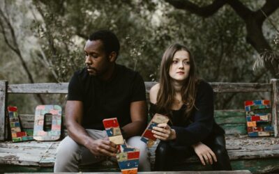 A couple sits in tension on a bench surrounded by nature, holding colorful blocks spelling 'Love'.