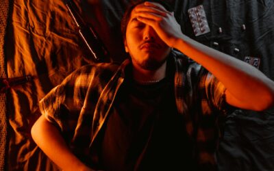 An adult man lies indoors with his hand on his face surrounded by pills and a bottle, indicating stress.