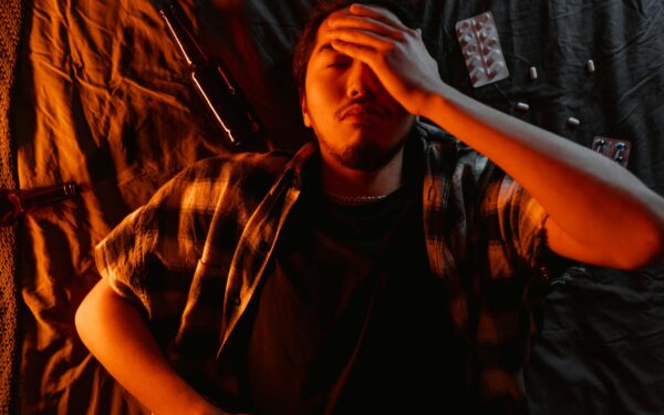 An adult man lies indoors with his hand on his face surrounded by pills and a bottle, indicating stress.