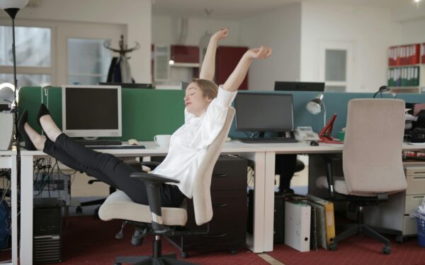 Side view of cheerful female employee in formal outfit sitting on office chair with crossed legs on desk and stretching while resting during work with closed eyes