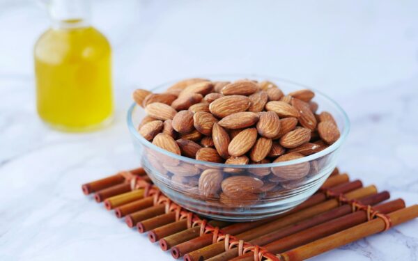 Close-up of almonds in a glass bowl next to a bottle of oil on a bamboo mat.