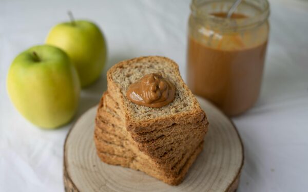 Stack of whole grain bread with peanut butter, accompanied by apples and a jar of smooth peanut butter.