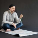 Man in traditional attire meditates on a prayer mat with beads, in a studio setting.