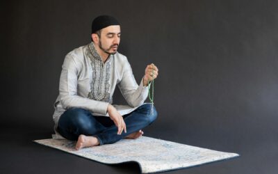 Man in traditional attire meditates on a prayer mat with beads, in a studio setting.