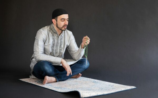 Man in traditional attire meditates on a prayer mat with beads, in a studio setting.