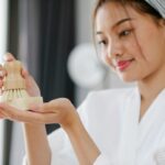 Charming Asian female in white bathrobe with soap and body brush for skincare treatment standing in room on blurred background