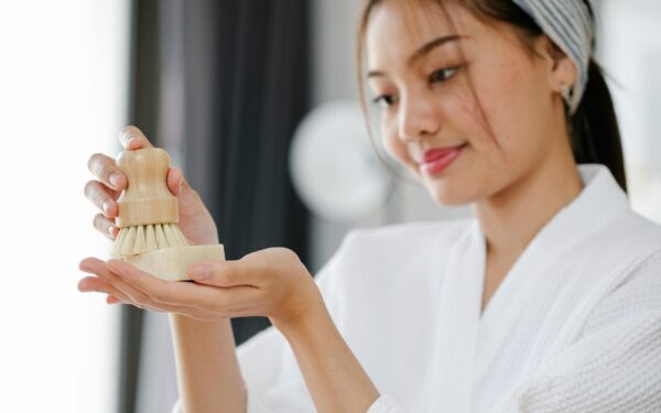 Charming Asian female in white bathrobe with soap and body brush for skincare treatment standing in room on blurred background