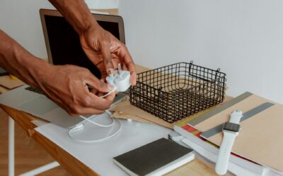 Close-up of a man organizing a desk, handling a charger next to a laptop, wire basket, and notebooks.
